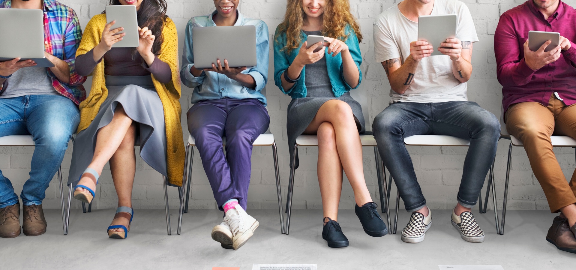 A group of smiling people sitting in a waiting room with electronic devices in each of their hands